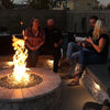 A lively evening gathering around a round stone fire pit filled with sparkling Azuria reflective fire glass. The tall, radiant flame creates a warm glow as friends laugh and chat on built-in stone seating, enjoying drinks and quality time together. Perfectly staged in a cozy backyard with soft lighting and privacy fencing—this is outdoor entertaining at its best, featured on starfiredirecv.com.