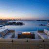 A large U-shaped sectional sofa surrounds a rectangular outdoor fire pit in a graphite finish, set against a backdrop of a harbor and city lights during blue hour. The low-profile fire table provides a warm gathering spot on the rooftop deck. Clean-burning flames illustrate the benefits of a smokeless fire pit, ensuring unobstructed views of the water while creating an inviting atmosphere for a family gathering fire pit setup.
