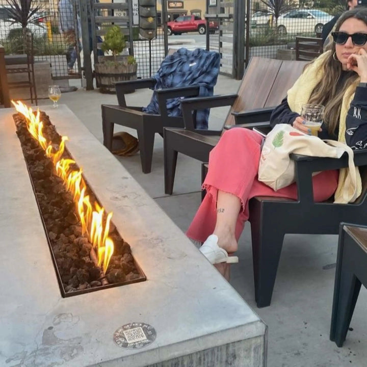Contemporary lounge featuring a long linear fire pit lined with decorative glass media and surrounded by industrial-style metal framework, fueled by the HPC Fire Inspired Linear Interlink Torpedo T-Burner.