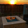 Modern square fire pit with turquoise fire glass ignited at sunset, set against a dark wood privacy fence, creating a warm and inviting ambiance for outdoor relaxation with HPC’s square burner technology.
