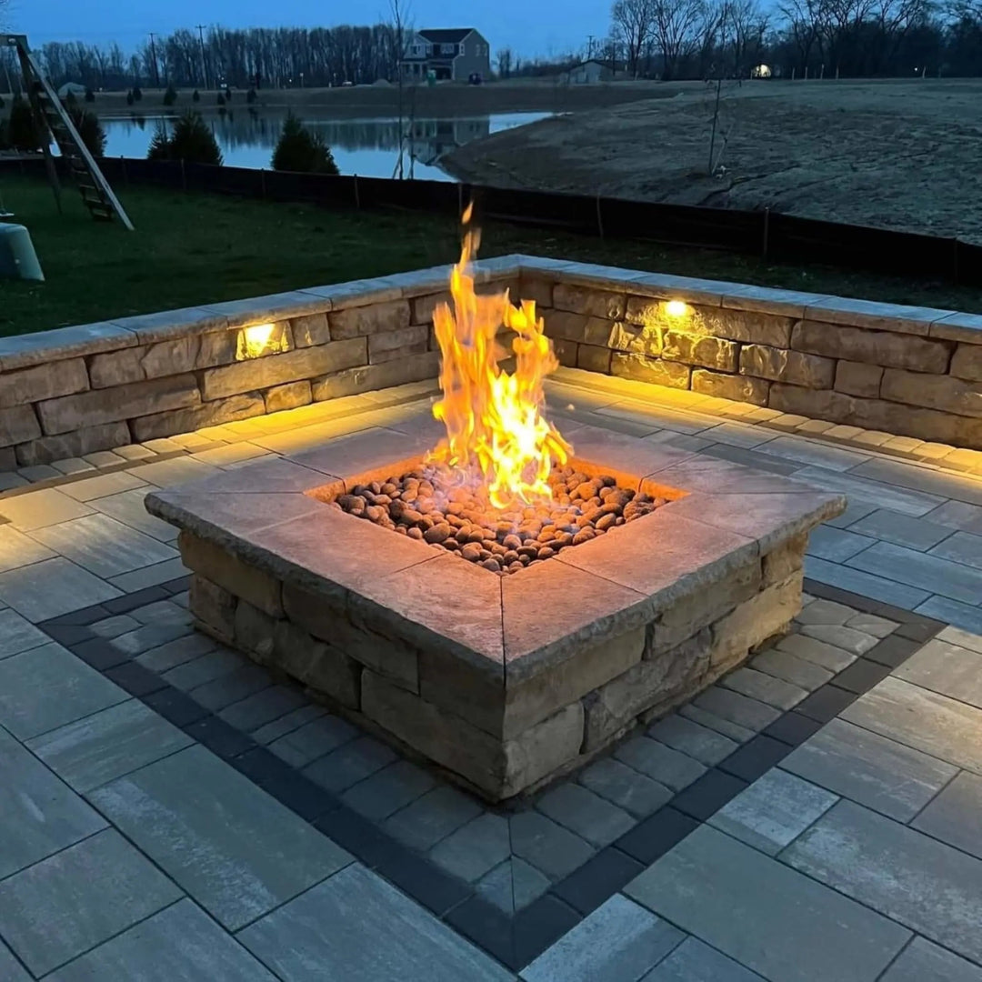 Square stone fire pit ignited with bright, vertical flames at dusk, surrounded by built-in stone seating and soft landscape lighting / an ideal backyard retreat powered by HPC's square flat burner system.