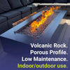 Modern fire table with glass wind guard showcasing bright flames over small gray lava stones—volcanic rock media ideal for indoor or outdoor use, offering a porous, low-maintenance design.