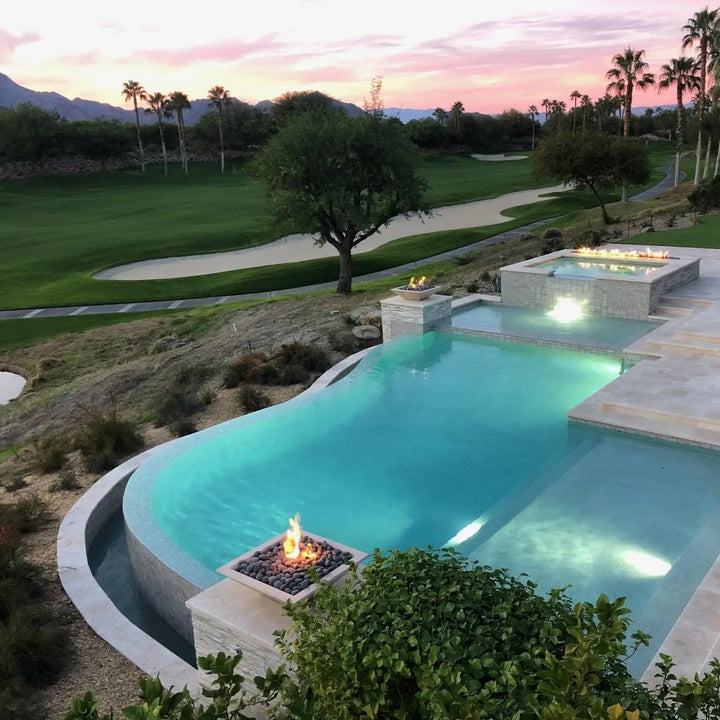 A breathtaking infinity-edge pool overlooking a golf course, featuring Maya Fire Bowls in Vanilla finish by The Outdoor Plus. The warm flames reflect against the water as palm trees and mountains complete the tranquil backdrop.