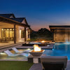 A picturesque backyard view at dusk with Maya Fire Bowls in Vanilla finish from The Outdoor Plus, installed along the edge of a glowing modern pool. The flickering fire elements add elegance and warmth to this beautifully lit luxury setting.