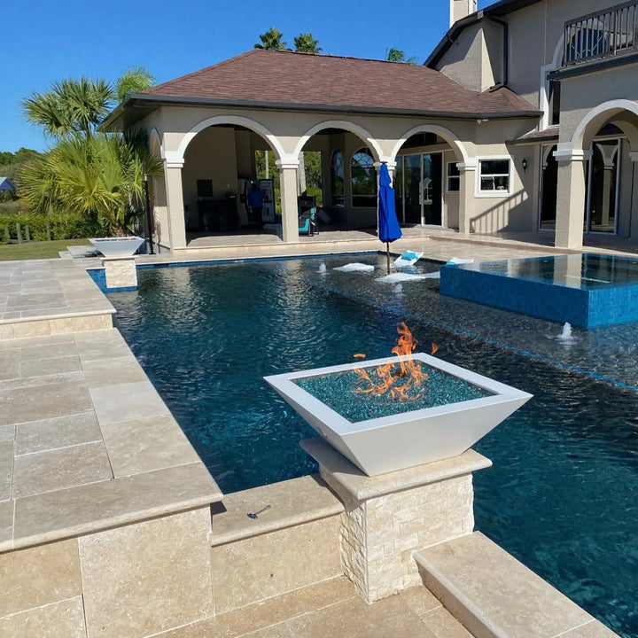 A breathtaking infinity-edge pool overlooking a golf course, featuring Maya Fire Bowls in Vanilla finish by The Outdoor Plus. The warm flames reflect against the water as palm trees and mountains complete the tranquil backdrop.