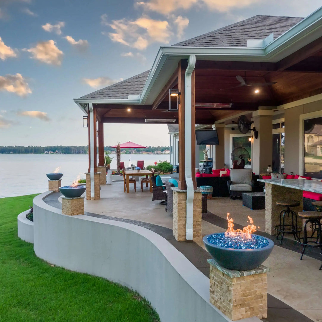 Gray Sedona Fire Bowls by The Outdoor Plus with blue fire glass, neatly positioned on stone columns across a lakefront patio, delivering breathtaking views and glowing warmth.
