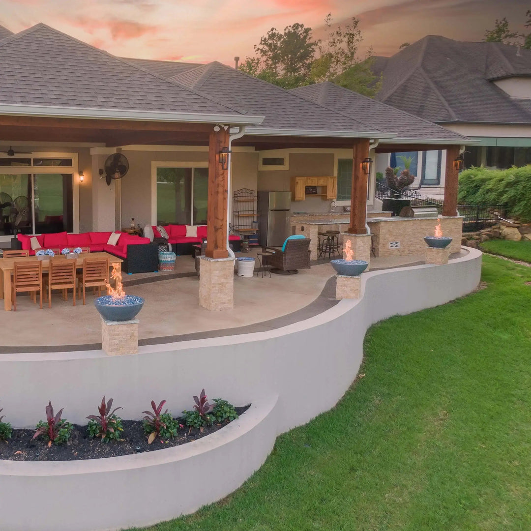 Gray Sedona Fire Bowls by The Outdoor Plus with blue fire glass, neatly positioned on stone columns across a lakefront patio, delivering breathtaking views and glowing warmth.