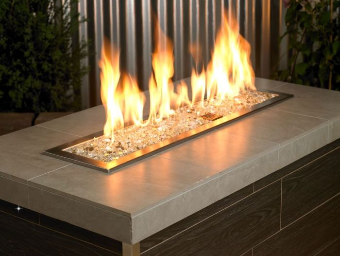 A sleek outdoor fire table glows warmly with golden flames dancing over a bed of 1/4" Gold Reflective Fire Glass by American Fire Products, adding luxury and sparkle to any modern patio or entertaining space.