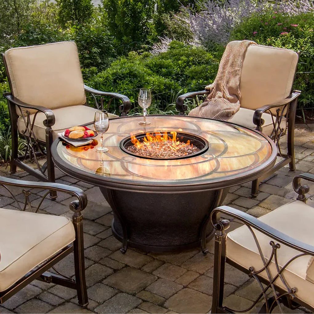 Elegant outdoor dining area with a round stone fire pit table and four cushioned wrought iron armchairs, set on a stone-paved patio surrounded by lush greenery, creating a warm and inviting ambiance for outdoor gatherings