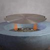 Starfire Designs Beton Round Stainless Steel Burner Cover being placed onto a fire pit, demonstrating the correct installation process with arrows indicating placement