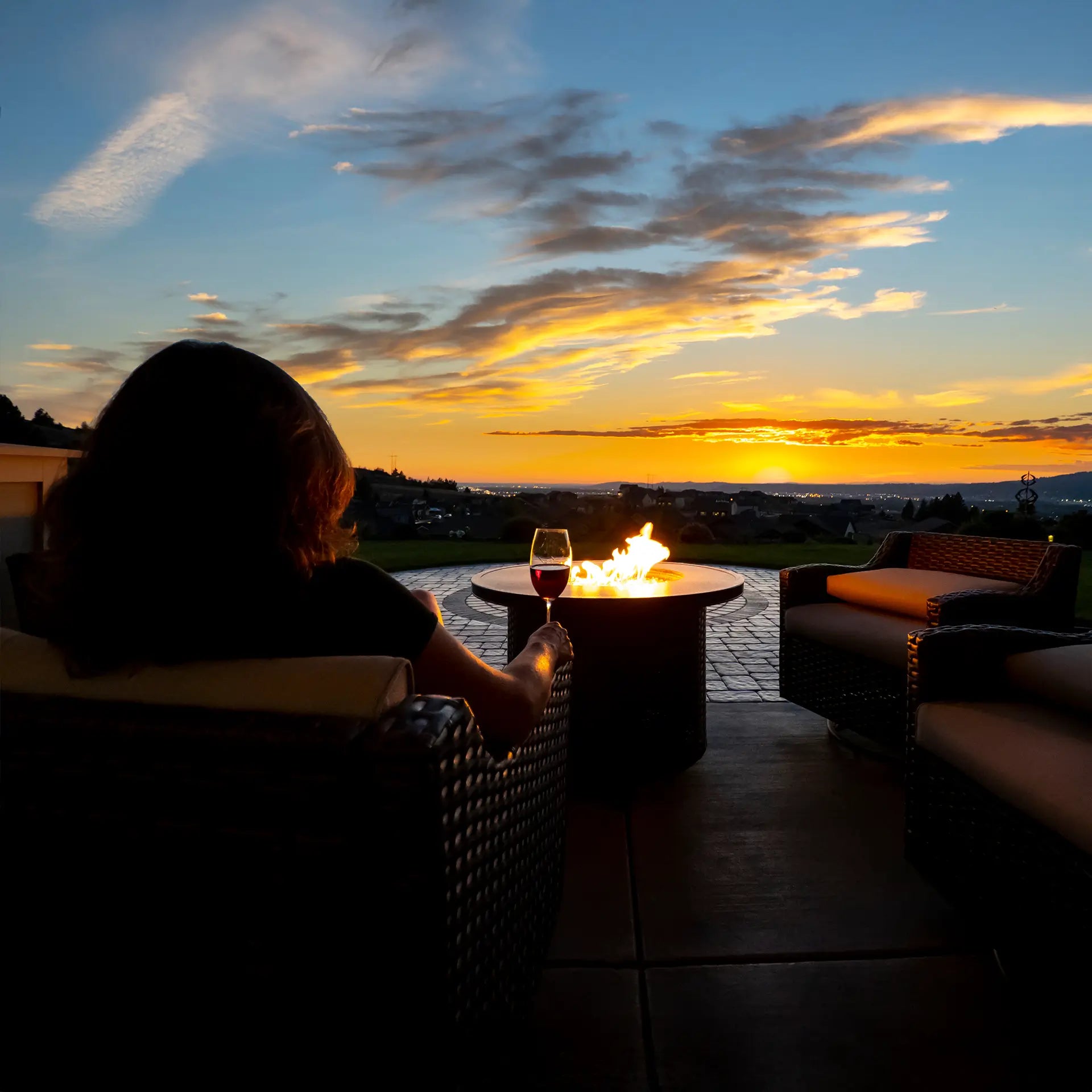 A woman sits in a comfortable patio lounge chair in front of a fire pit table drinking wine while watching the sunset in her own backyard.
