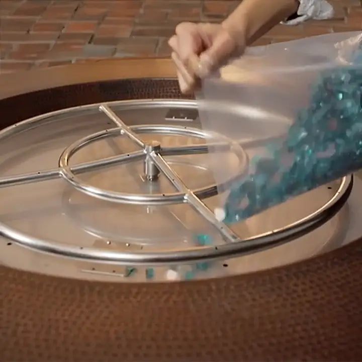 A woman pours out fire glass onto a burner pan in a fire pit to decorate it.