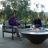 Cono Moreno 42-inch Round Copper Fire Pit with a hand-hammered finish, surrounded by modern patio seating, perfect for an outdoor gathering. Two people enjoy a cozy evening with wine around the warm, inviting glow of the fire pit, featuring blue fire glass