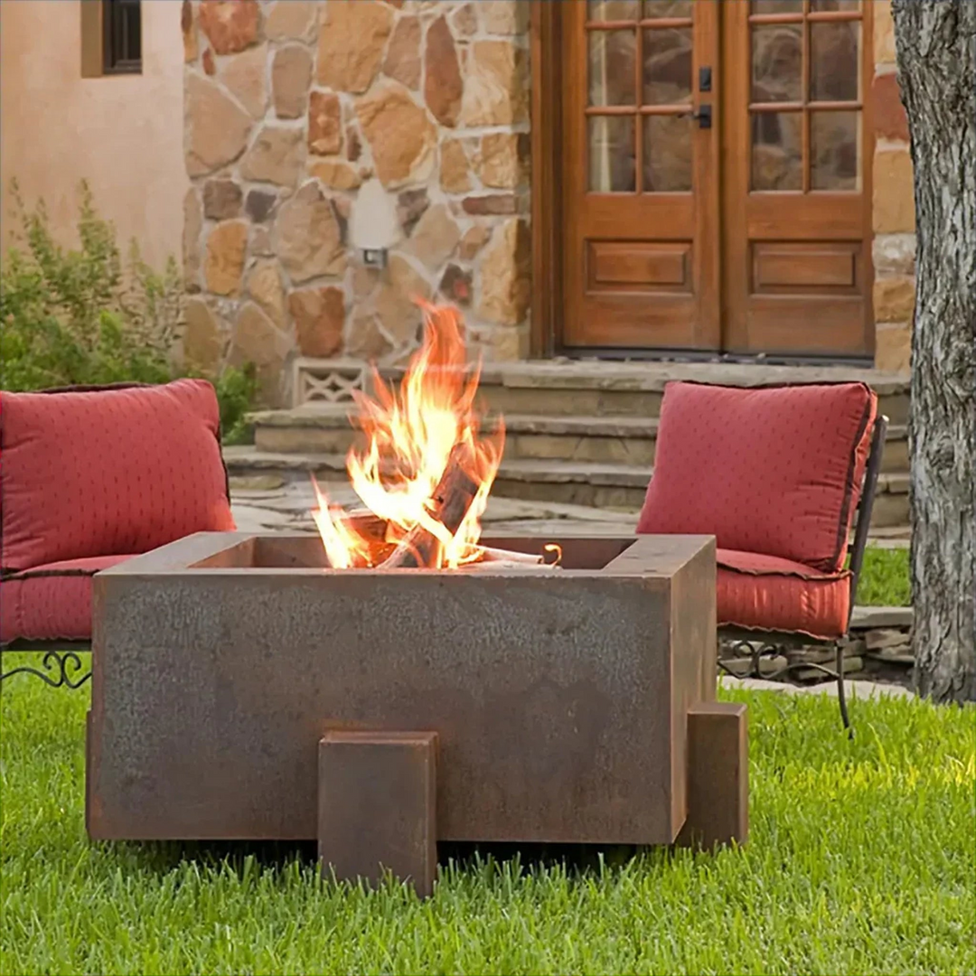 Rustic square fire pit in a backyard setting with a lit fire, surrounded by red cushioned chairs and a stone house in the background