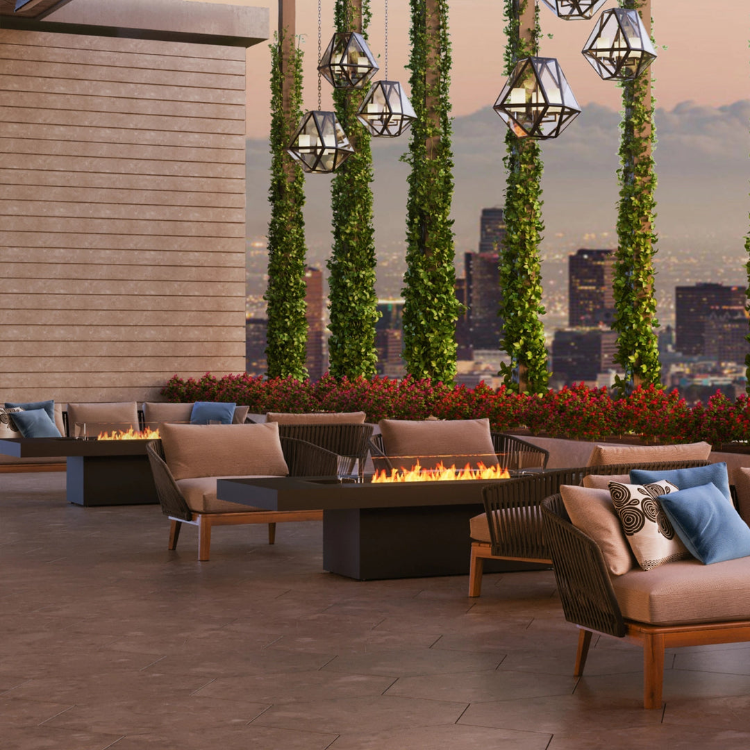 The EcoSmart Fire Gin 90 Chat Height fire pit table in a sleek graphite finish, elegantly placed on a luxurious rooftop terrace with plush seating and stunning skyline views. The rectangular gas fire table provides a warm ambiance, making it the perfect centerpiece for an outdoor lounge. This modern outdoor fire pit features a low-profile design, glass wind guard, and clean-burning fire for a stylish and functional outdoor space.