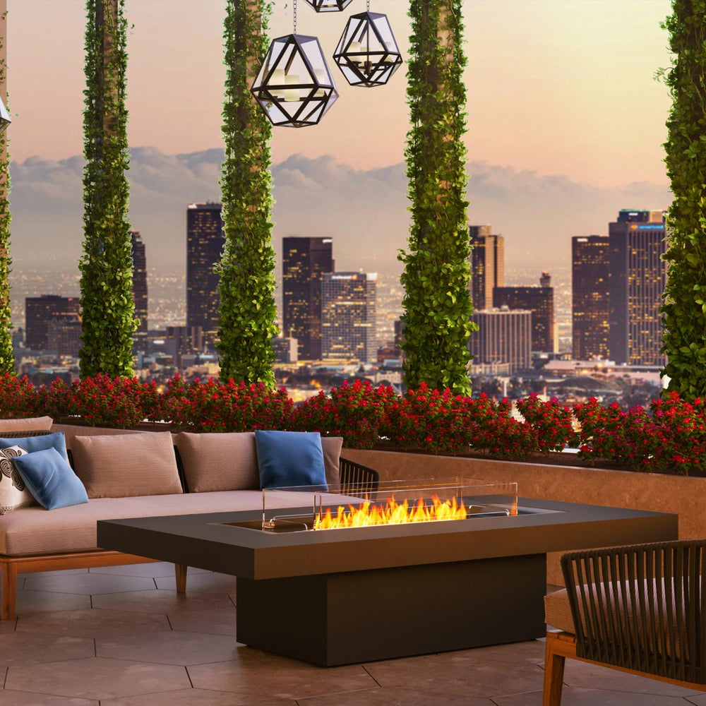 The EcoSmart Fire Gin 90 Chat Height outdoor fire table enhances a contemporary rooftop lounge, featuring comfortable patio seating, decorative lighting, and lush greenery. This rectangular gas fire pit with a glass wind guard offers a luxurious atmosphere for entertaining. Designed with high-quality materials, this modern fire table is ideal for commercial patios, high-end outdoor lounges, and backyard spaces.