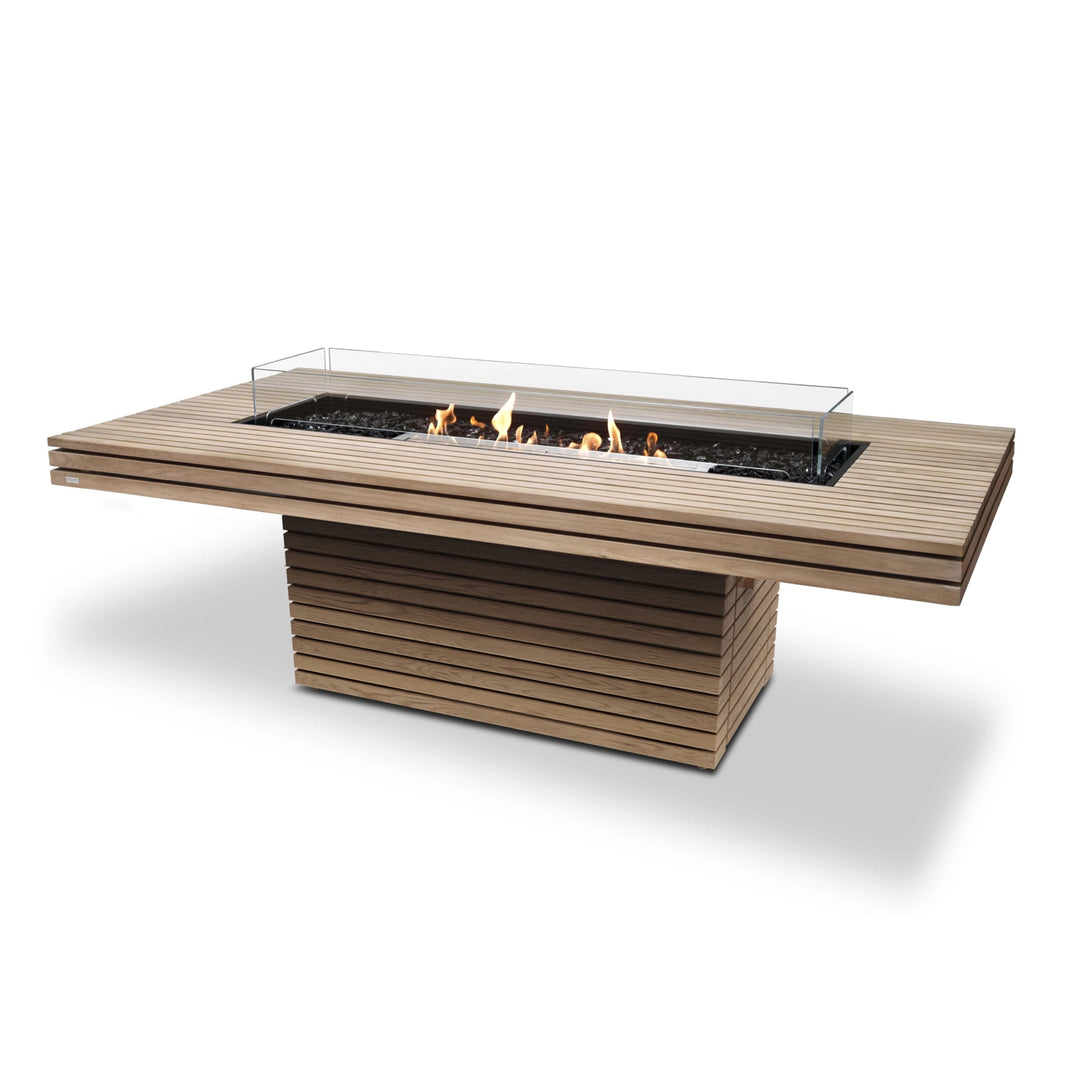 This EcoSmart Fire Gin 90 Dining Height Teak outdoor fire table showcases a modern teakwood finish with horizontal slats and a sophisticated glass wind guard. The gas fire bowl provides consistent warmth with a stainless steel burner, making it an excellent choice for those seeking a high-end fire pit for entertaining in commercial fire pit settings or luxury outdoor spaces.