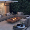 The EcoSmart Fire Gin 90 Low fire table in a cozy outdoor lounge area at dusk. Featuring teak wood slats and a glass wind guard, this modern fire pit provides a sophisticated focal point among plush sectionals. The warm natural gas flames add a cozy touch, ideal for backyard entertaining and outdoor relaxation.