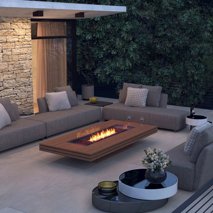 The EcoSmart Fire Gin 90 Low fire table in a cozy outdoor lounge area at dusk. Featuring teak wood slats and a glass wind guard, this modern fire pit provides a sophisticated focal point among plush sectionals. The warm natural gas flames add a cozy touch, ideal for backyard entertaining and outdoor relaxation.