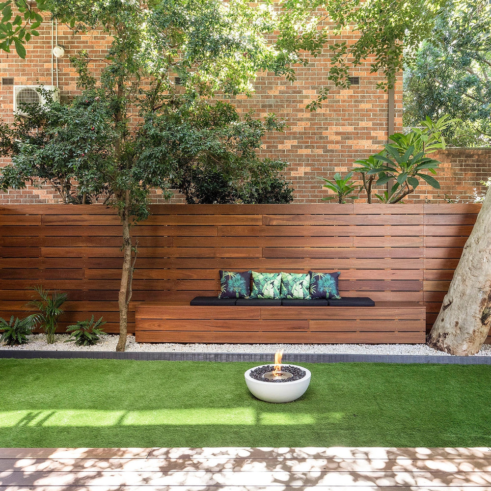 The EcoSmart Fire Mix 600 Bone Fire Pit is featured in an elegant outdoor patio setting, placed on a lush green lawn with a wooden bench and decorative pillows in the background. This modern fire pit bowl is filled with black lava rocks and emits a warm, inviting flame, making it the perfect centerpiece for any backyard fire pit arrangement. Designed for both aesthetics and functionality, it provides an eco-friendly fire pit outdoor experience.