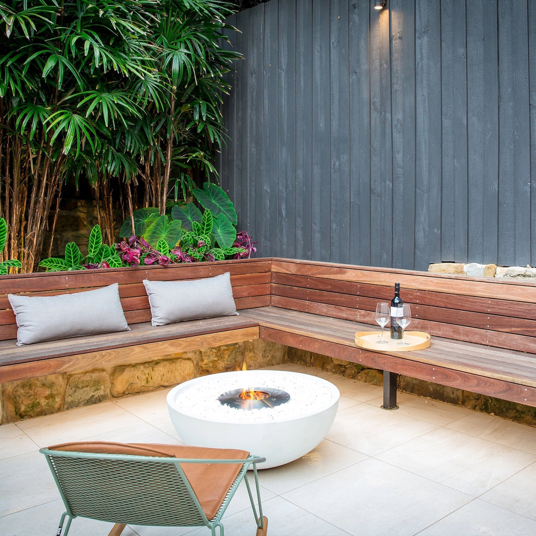 The EcoSmart Fire Mix 850 bone fire pit is beautifully integrated into a cozy garden seating area. The elegant round fire bowl is filled with white fire media, complementing the lush greenery and wooden bench seating. This outdoor gas fire pit creates a warm and intimate atmosphere, perfect for backyard patios and lounge spaces.

