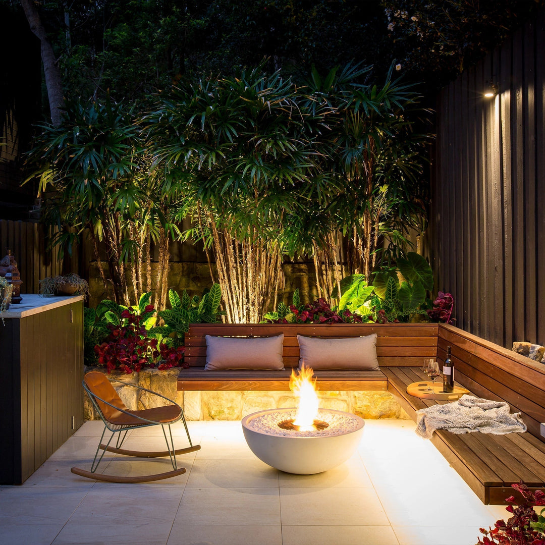 The EcoSmart Fire Mix 850 bone fire pit is the centerpiece of an elegant outdoor lounge area, surrounded by lush plants and ambient lighting. Its round concrete fire bowl with white fire glass enhances the inviting atmosphere, making it ideal for modern backyard fire pit setups and patio heating solutions.