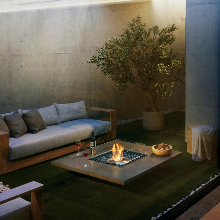 EcoSmart Fire Vertigo 50 lifestyle setting featuring a sleek modern outdoor fire pit with a natural concrete finish, surrounded by a cozy seating arrangement with a wooden sofa and armchair. The fire pitâ€™s clean lines, glass wind guard, and glowing flames create a stylish and functional focal point for any contemporary patio or backyard space.