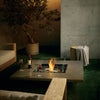  EcoSmart Fire Vertigo 50 showcased in a nighttime outdoor setting, creating a warm and intimate ambiance. The fire pitâ€™s sleek natural finish, glass wind guard, and radiant flames complement the modern patio dÃ©cor, featuring a minimalist wooden sofa, armchairs, and a potted olive tree. Glasses of red wine on the table enhance the cozy, luxurious atmosphere