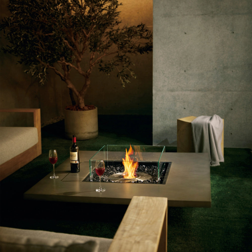  EcoSmart Fire Vertigo 50 showcased in a nighttime outdoor setting, creating a warm and intimate ambiance. The fire pitâ€™s sleek natural finish, glass wind guard, and radiant flames complement the modern patio dÃ©cor, featuring a minimalist wooden sofa, armchairs, and a potted olive tree. Glasses of red wine on the table enhance the cozy, luxurious atmosphere