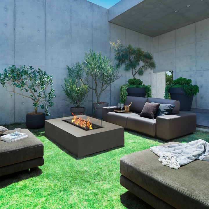 A serene minimalist courtyard with lush greenery, showcasing the EcoSmart Fire Wharf 50 in a modern dark concrete finish. The large outdoor fire pit seamlessly blends into the contemporary landscape, offering a sophisticated luxury fire pit experience for both residential and commercial outdoor spaces.