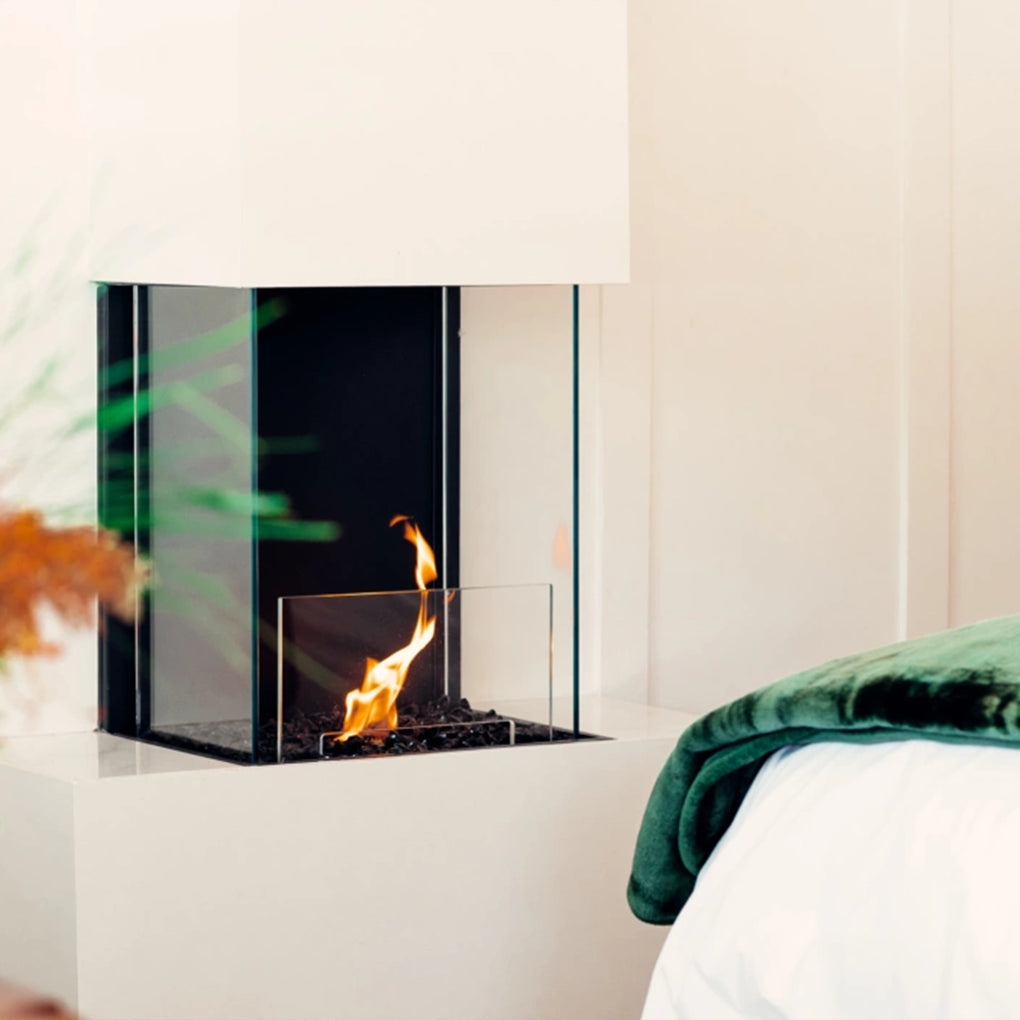 Close-up of a modern corner fireplace with a glass enclosure, integrated into a minimalist white wall, adjacent to a cozy bed with a dark green blanket, creating a warm and inviting bedroom setting