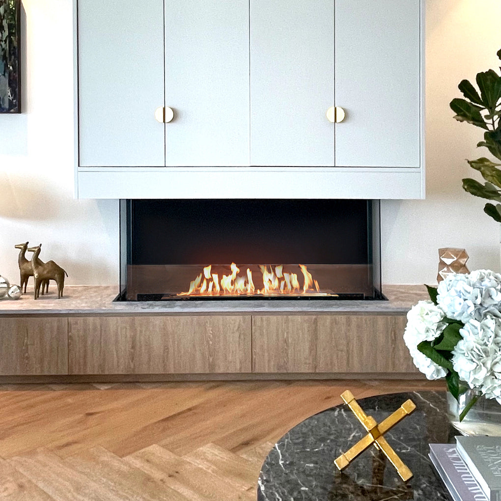 Stylish modern living room featuring a sleek linear fireplace below white cabinets with brass knobs, light wood accents, marble table with a gold decorative piece, brass animal figurines, and a fresh floral arrangement, creating an elegant and contemporary atmosphere