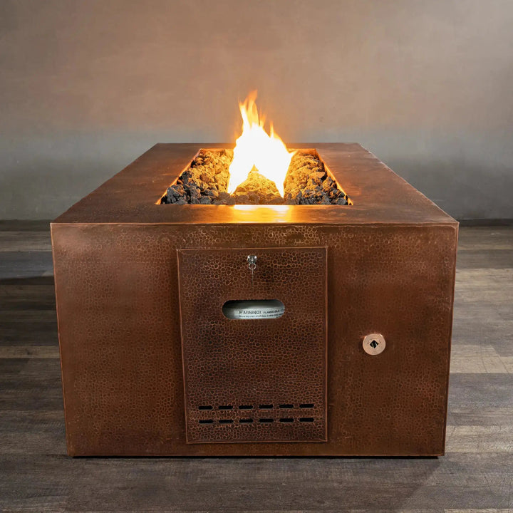Close-up view of the Gravity Moreno 72x38 copper fire pit by Starfire Designs, featuring the propane tank access door with a warm, inviting flame in the background