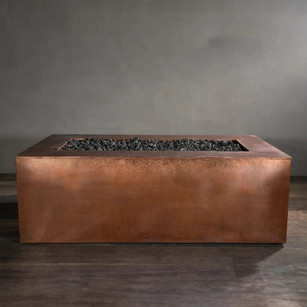 Side view of the Gravity Moreno 72x38 copper fire pit by Starfire Designs, showcasing its elegant hammered copper finish and spacious fire bed filled with black lava rocks