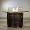 The Golden Flame Malibu 44” Round Cast Aluminum Natural Gas Fire Table, showcasing its elegant bronze finish and intricate pattern with flames glowing beautifully in a glass wind guard.
