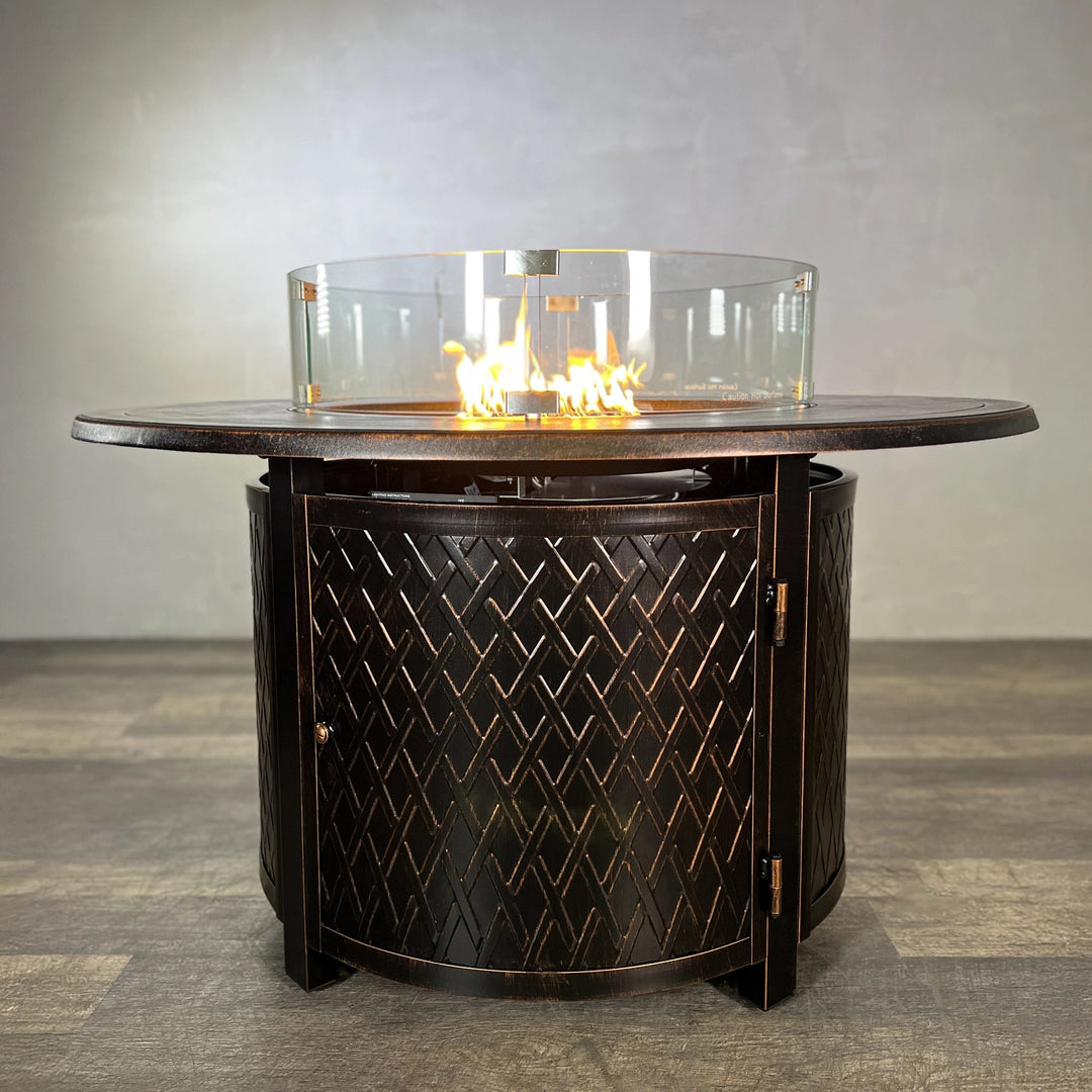 The Golden Flame Malibu 44” Round Cast Aluminum Natural Gas Fire Table, showcasing its elegant bronze finish and intricate pattern with flames glowing beautifully in a glass wind guard.