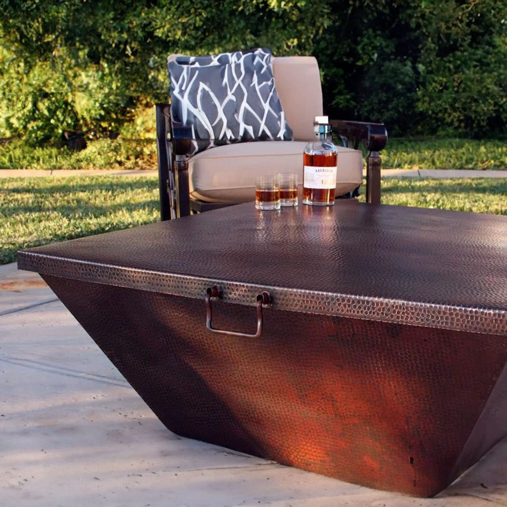 Moreno 40-inch x 40-inch square copper fire pit lid in an outdoor setting, placed over a copper fire pit by Starfire Designs. This hammered copper lid is built to withstand outdoor conditions, offering both protection and style to your fire pit setup. Perfect for large outdoor fire bowls, backyard fire pits, and patio fire tables, the lid features a natural copper finish and a sturdy handle for convenience