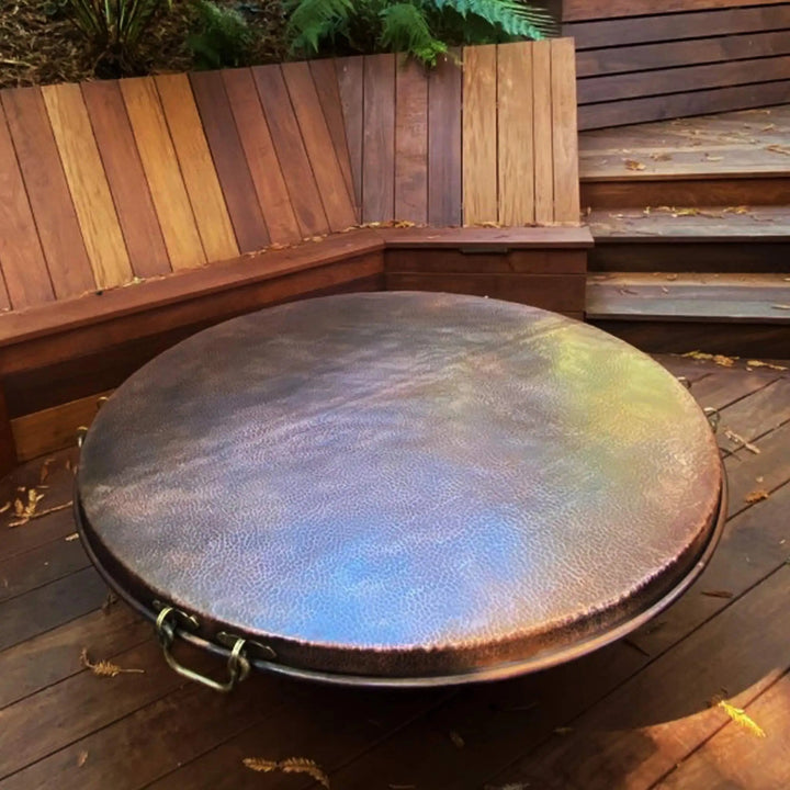 This image showcases the Moreno 42" round copper fire pit lid placed on a patio, highlighting its beautiful hammered copper texture. The fire pit cover is designed to protect the fire pit while doubling as an attractive tabletop. The handcrafted copper finish adds luxury to any outdoor setup. Ideal for outdoor fire pits, this round copper lid features durable handles for easy removal and maintenance.