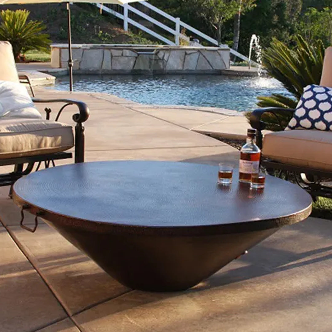 Modern outdoor fire pit bowl repurposed as a table with a whiskey bottle and glasses, surrounded by patio furniture in a poolside setting with a fountain and stone retaining wall.