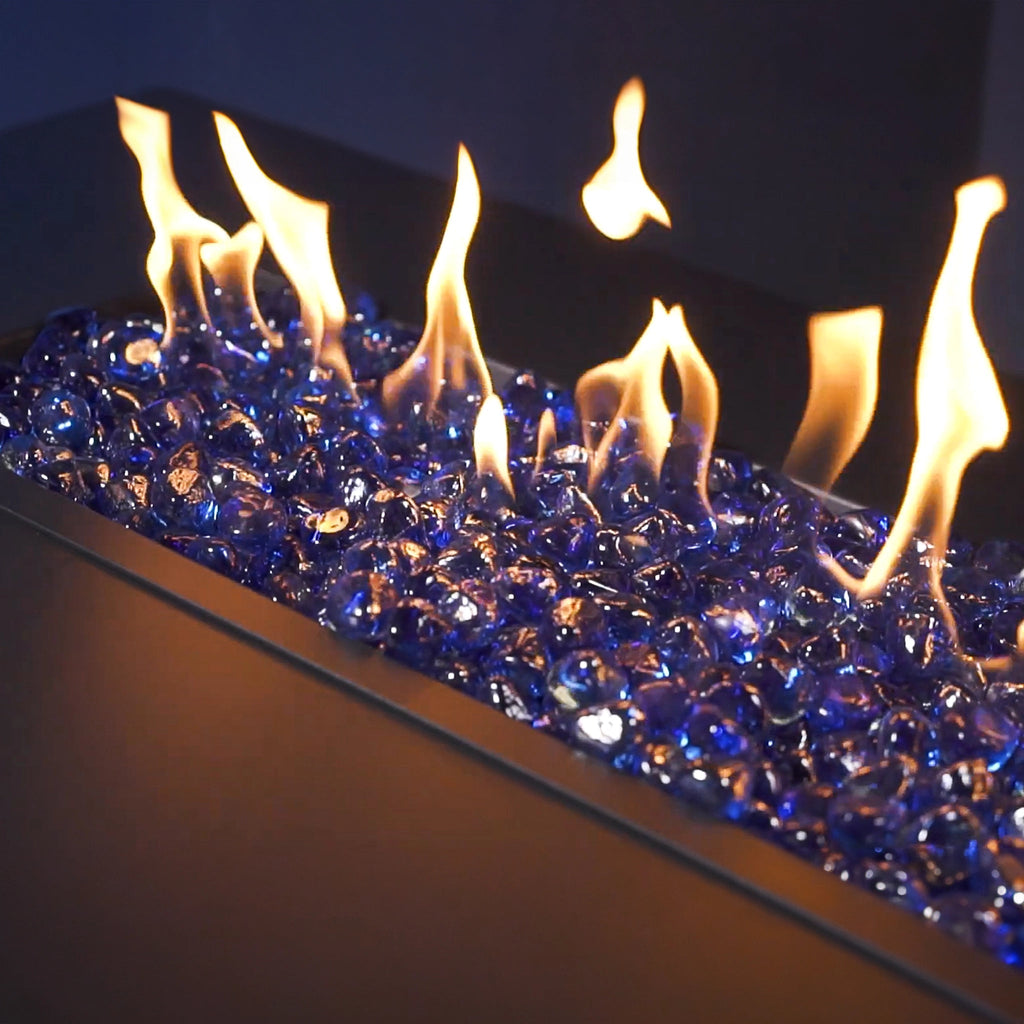 Mr. Patio Fire Glass Fire-Diamonds glowing in a gas fire pit, with vibrant blue glass stones shimmering under the bright flames. The combination of fire and glass creates a mesmerizing effect, making the fire pit a focal point for outdoor entertaining. Designed for durability and beauty, the fire diamonds are made from high-quality tempered glass that can withstand extreme heat and is suitable for both indoor and outdoor use. These blue fire diamonds are ideal for enhancing the ambiance of any fire feature