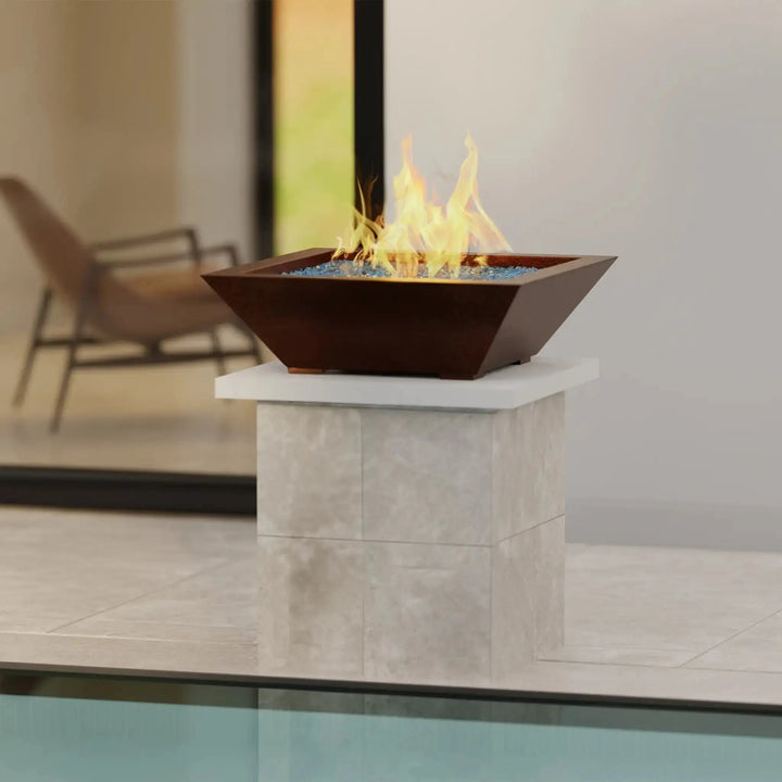 Plaza Moreno 29-inch square copper fire bowl on a modern outdoor pedestal, showcasing a natural gas flame with blue fire glass, ideal for contemporary patio designs
