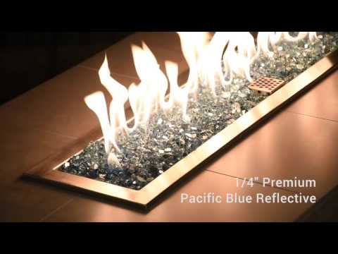 Vibrant Pacific Blue 1/4" Reflective Fire Glass from American Fire Products glowing beneath bright flames in a modern fire feature—its rich ocean-blue tones and mirrored surfaces create a striking visual contrast, perfect for enhancing outdoor ambiance with color and shine.