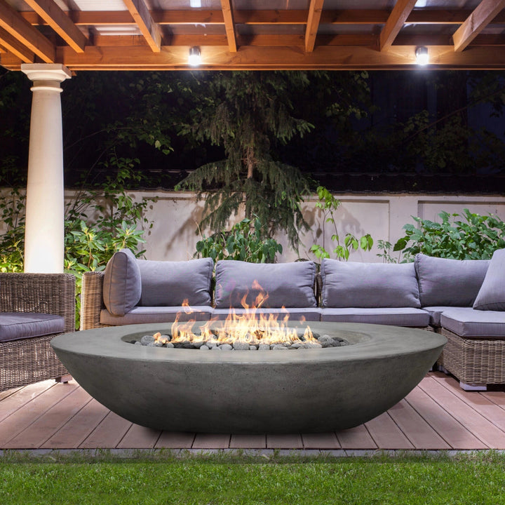 Prism Hardscapes Ovale Gas Fire Pit showcased in a luxurious backyard setting, surrounded by comfortable patio furniture under a wooden pergola. The fire pit features a smooth, round, modern design in natural concrete, providing a stunning focal point during an evening gathering. Its warm flames illuminate the outdoor space, creating a cozy and inviting atmosphere, perfect for modern fire pits, outdoor gas fire pits, and luxury backyard settings
