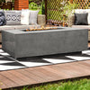 The Prism Hardscapes Portos 58 Propane Fire Table in gray finish is displayed in a cozy evening patio setting, surrounded by lush greenery and comfortable wicker furniture with gray cushions. The rectangular propane fire table emits a warm flame, perfect for relaxing or entertaining guests outdoors. Set on a wooden deck, this fire table provides an elegant modern outdoor fire feature that complements the natural surroundings, enhancing the ambiance