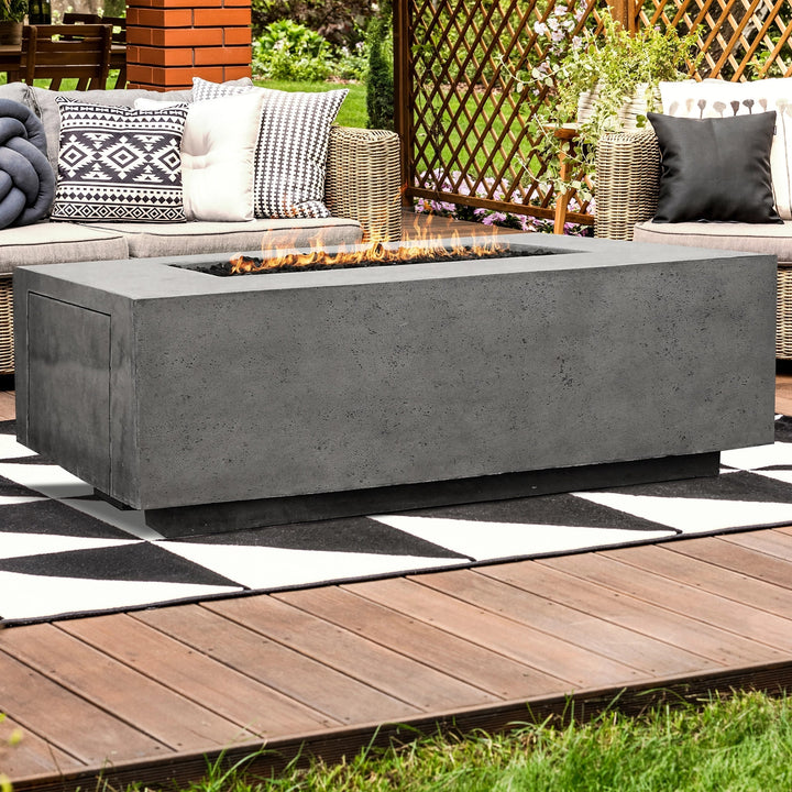 The Prism Hardscapes Portos 58 Propane Fire Table in gray finish is displayed in a cozy evening patio setting, surrounded by lush greenery and comfortable wicker furniture with gray cushions. The rectangular propane fire table emits a warm flame, perfect for relaxing or entertaining guests outdoors. Set on a wooden deck, this fire table provides an elegant modern outdoor fire feature that complements the natural surroundings, enhancing the ambiance