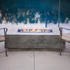 Prism Hardscapes Tavola 1 Gas Fire Pit in natural gray, elegantly placed poolside with a backdrop of water. The sleek rectangular fire pit features a large burner with stunning blue fire glass, providing a luxurious and modern outdoor fire pit experience. Ideal for poolside patios, this natural gas fire pit creates warmth and ambiance, perfect for outdoor entertaining.