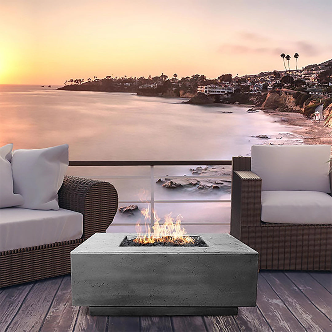 A stunning Prism Hardscapes Tavola 3 Gas Fire Pit is featured on a deck overlooking a breathtaking sunset ocean view. This modern concrete gas fire pit creates a cozy and inviting atmosphere, making it the perfect spot for relaxation. With wicker chairs and a clear glass barrier protecting the flame, this fire pit is ideal for coastal settings and adds a touch of luxury to any outdoor space. The propane-powered fire pit offers warmth and style, blending seamlessly into the serene environment. 