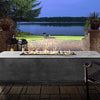 The Prism Hardscapes Tavola 4 Gas Fire Pit illuminates an evening patio setting, casting a warm glow over a scenic lakeside view. Its rectangular design and concrete finish provide a sophisticated touch to the patio space, making it the perfect addition for evening relaxation and outdoor entertainment. Ideal for those who enjoy the outdoors, this gas fire pit is both stylish and functional. 