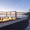 A serene lakeside evening with a lit Prism Hardscapes Tavola 6 Gas Fire Pit in the foreground. The fire pit's concrete structure adds to the tranquil setting, providing warmth as the sun sets over the water. Ideal for waterfront patios or decks, this fire pit offers a perfect blend of modern style and natural beauty.