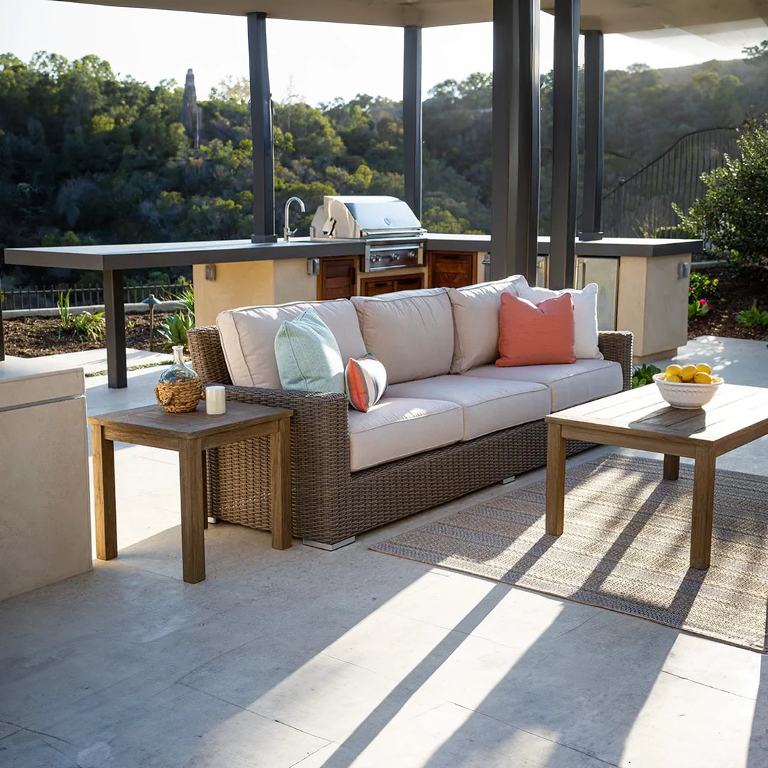 Outdoor patio setting featuring a wicker sofa with beige cushions, paired with a wooden coffee table and side table. The backdrop includes a built-in outdoor kitchen with a grill and a scenic landscape
